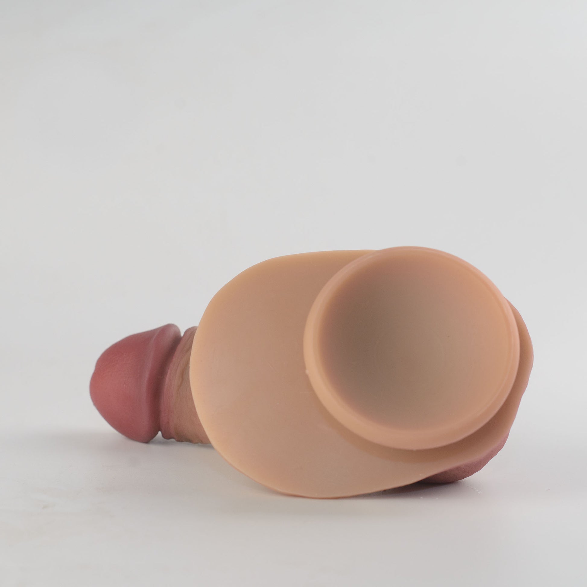 Stage 05 Ossifying Dragon dual-density silicone dildo - side-angled view of product lying flat showing suction cup base cavity, oversized scrotum and deep pink glans in background - 2.36in/6cm diameter suction cup for strong hands-free surface adhesion - body-safe platinum-cured | RealTouch Lab
