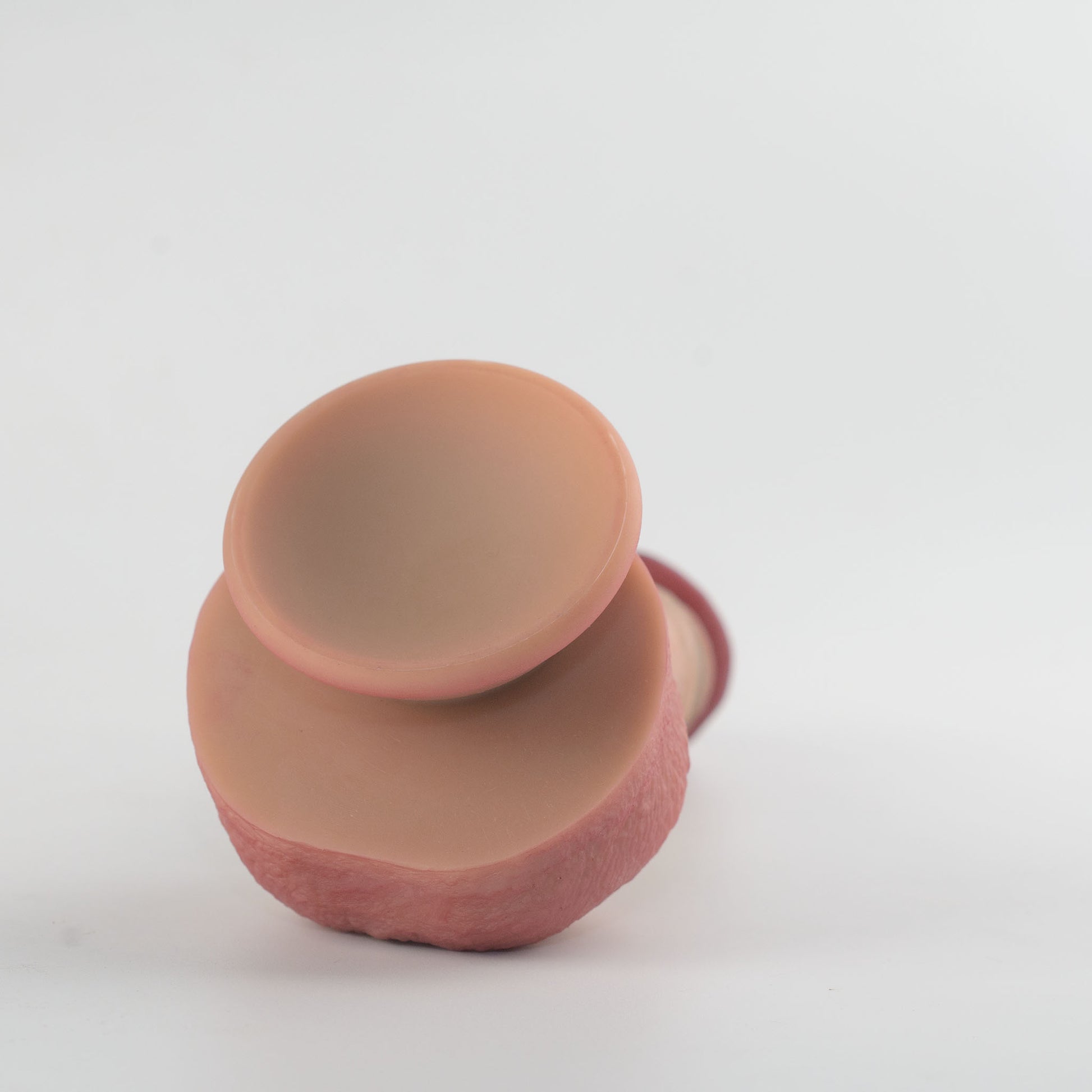 Stage 05 Velociraptor dual-density silicone dildo with Velvet Shield Coating - side-angled view of product lying flat showing smooth natural skin-tone suction cup base cavity and deep pink scrotum surround - 2.36in/6cm diameter suction cup for strong hands-free surface adhesion - body-safe platinum-cured | RealTouch Lab

