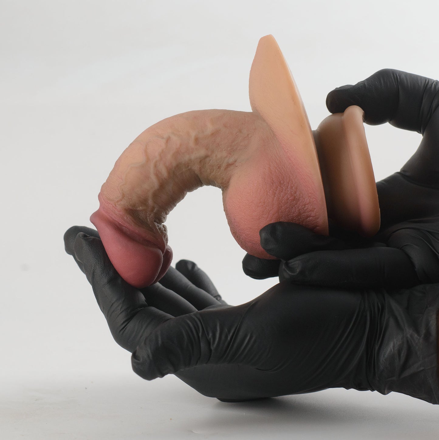 Stage 05 Ossifying Dragon dual-density silicone dildo - two black nitrile gloved hands bending product into U-shape demonstrating extreme dual-density flexibility, glans nearly touching scrotum with no surface cracking - oversized scrotum clearly visible under maximum bend - body-safe platinum-cured | RealTouch Lab
