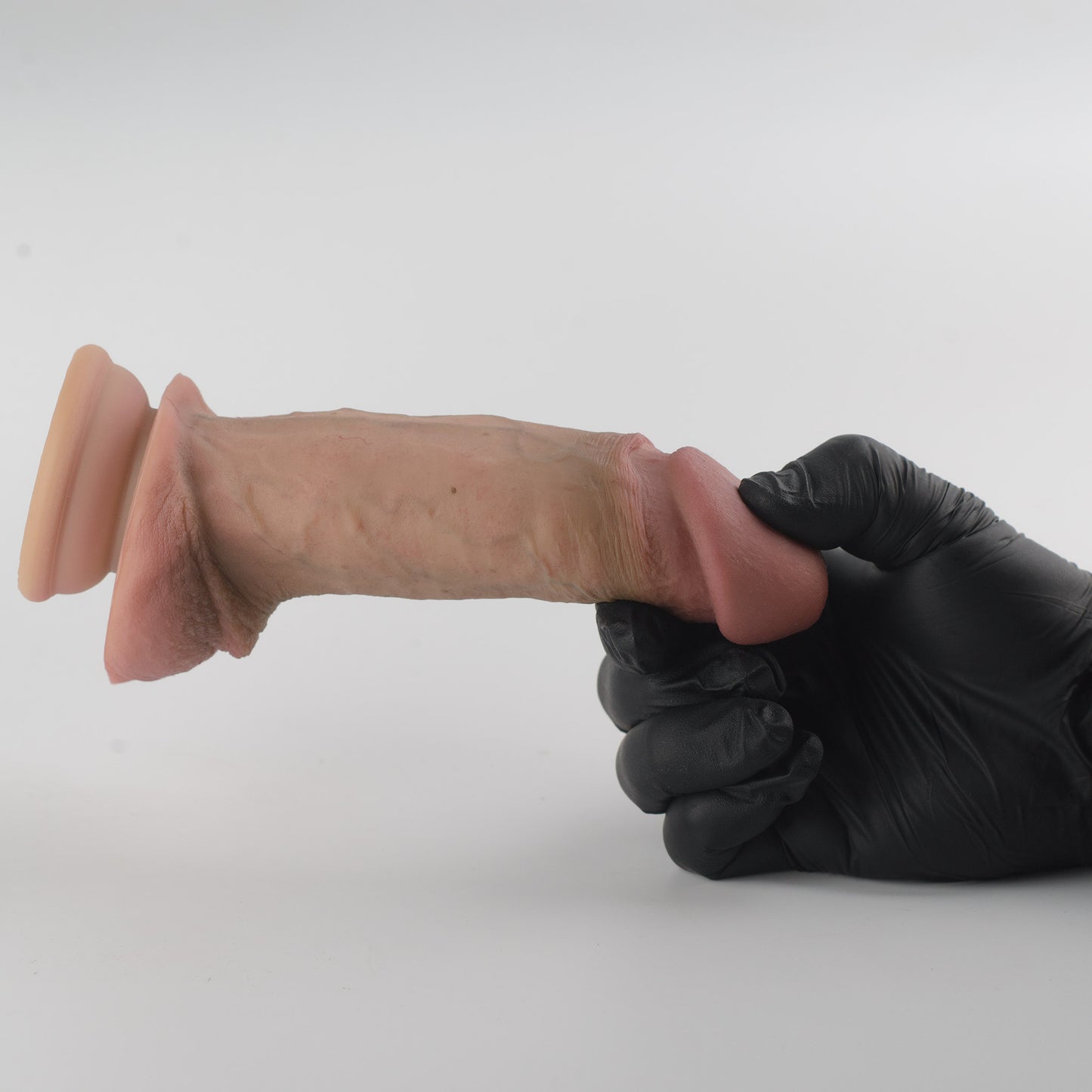 Stage 05 Brontosaurus dual-density silicone dildo with Velvet Shield Coating - black nitrile gloved hand gripping shaft near glans with product extended horizontally showing full side profile, deep pink glans, natural skin-tone shaft with vein detail, full scrotum and suction cup base on left - Velvet Shield Coating delivers matte finish and silky-smooth non-tacky surface - body-safe platinum-cured | RealTouch Lab
