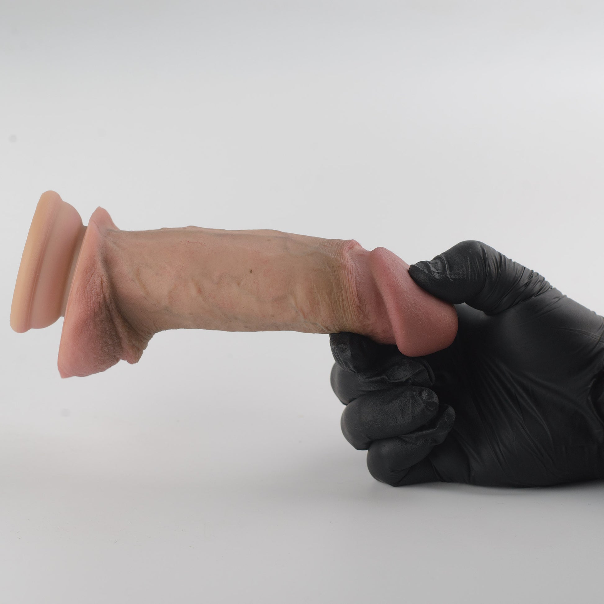 Stage 05 Brontosaurus dual-density silicone dildo with Velvet Shield Coating - black nitrile gloved hand gripping shaft near glans with product extended horizontally showing full side profile, deep pink glans, natural skin-tone shaft with vein detail, full scrotum and suction cup base on left - Velvet Shield Coating delivers matte finish and silky-smooth non-tacky surface - body-safe platinum-cured | RealTouch Lab
