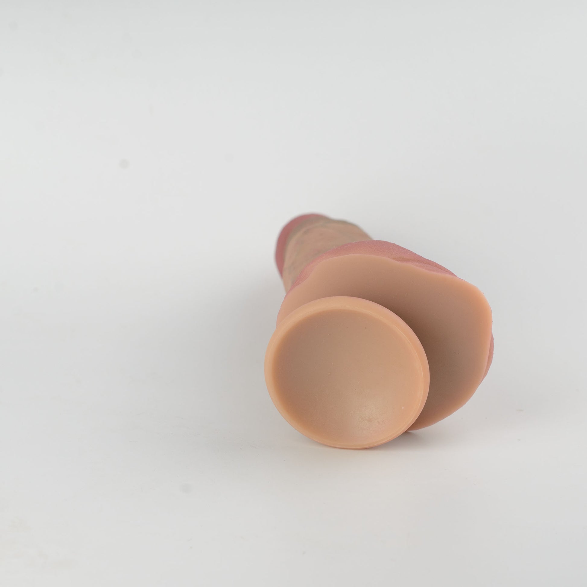 Stage 05 Brontosaurus dual-density silicone dildo with Velvet Shield Coating - side-angled view of product lying flat showing smooth natural skin-tone suction cup base cavity, full scrotum and deep pink glans visible in background - 2.36in/6cm diameter suction cup for strong hands-free surface adhesion - body-safe platinum-cured | RealTouch Lab
